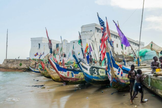 Colourful wooden fishing boats with flags lined up on a beach before a white coastal fort, a scenic stop on Treetops, Castles & Coastal Adventures day trips from Accra.