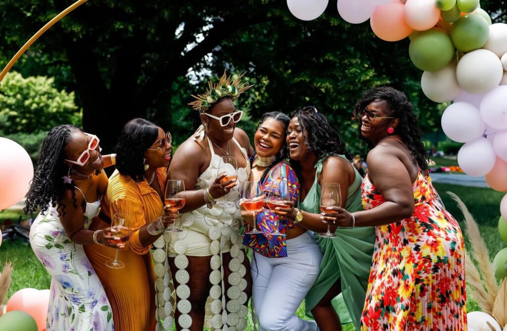 Six women laughing and holding glasses of rosé outdoors, with an arch of balloons in the background.