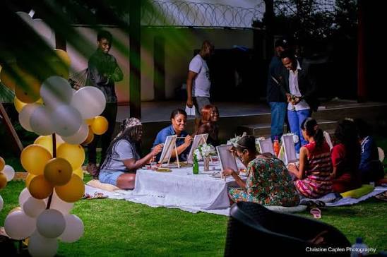 A group of people sitting at a table on a lawn, painting on canvases.