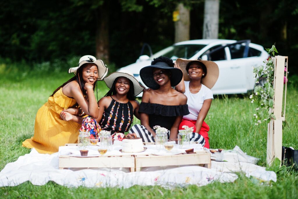 Four women wearing sun hats sitting on a blanket at an outdoor picnic with a cake and a white car in the background.